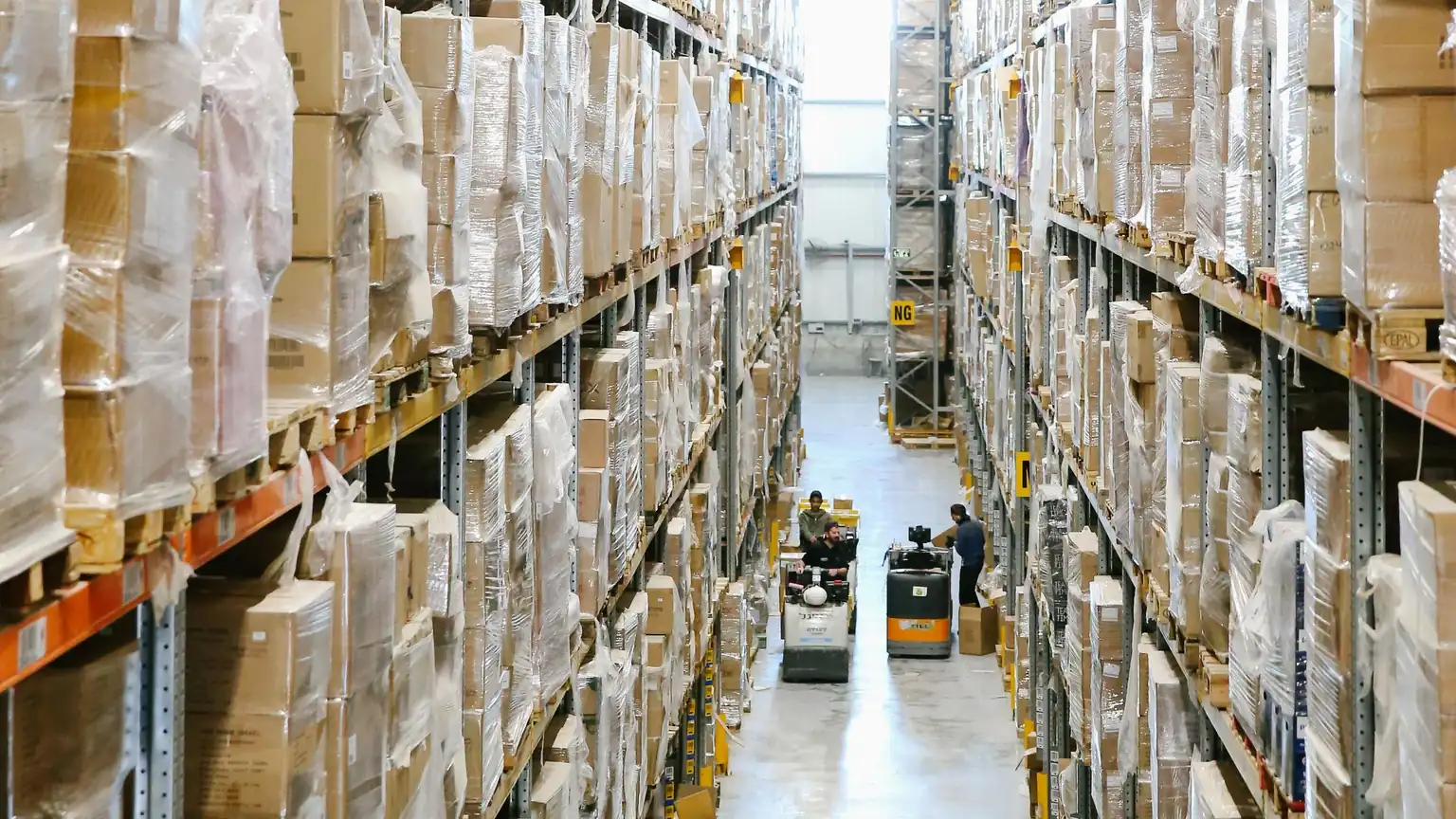 Lối đi giữa hai dãy kệ kho cao chất đầy thùng hàng trên pallet, hai xe nâng ở cuối lối