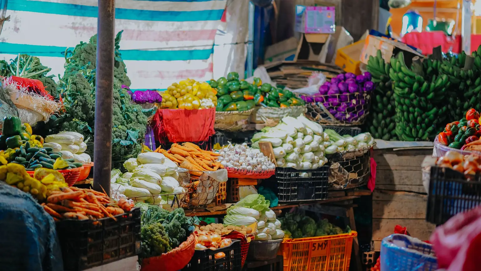 Sạp chợ đầy rổ rau củ tươi: cà rốt, bắp cải, súp lơ, chuối xếp trong két nhựa